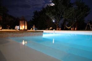 Una piscina con borde de azulejos, rodeada de árboles y mobiliario exterior.