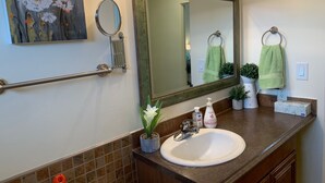 Ein Badezimmer mit einem weißen Waschbecken, einem großen Spiegel, einem Handtuchhalter mit grünen Handtüchern, einem kleinen Topfpflanzen und einem Blumenbild an der Wand.