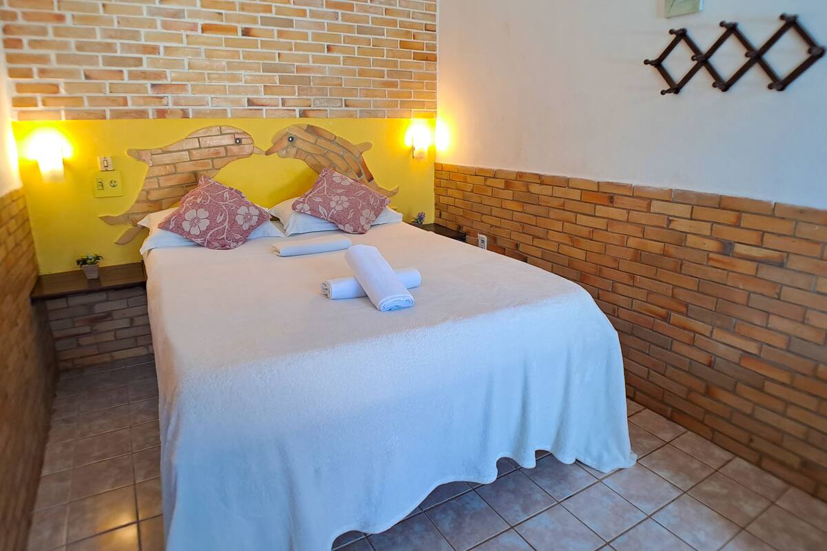Un lit bien fait, avec un couvre-lit blanc et des coussins à motifs floraux, un mur de briques et un mur jaune agrémenté d’une tête de lit décorative.