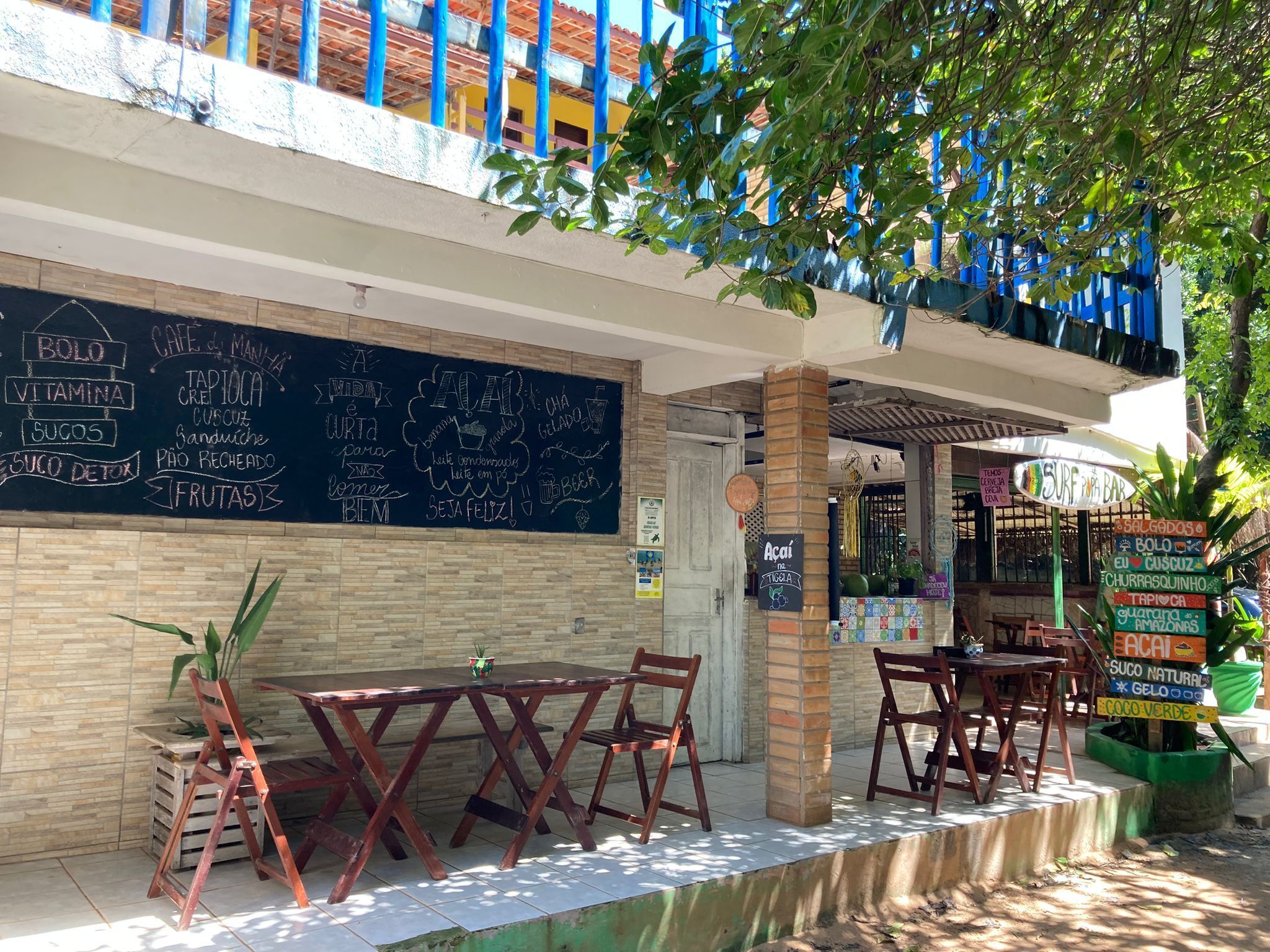 An outdoor café with wooden tables and chairs, a chalkboard menu, and a sign for a surf bar.