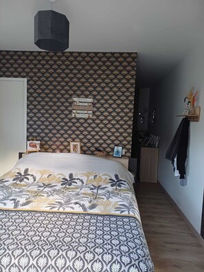 A bedroom with a patterned wallpaper, a bed with a floral bedspread, and a small shelf with decorative items.
