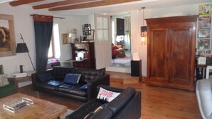 Un salón con un sofá de cuero negro, una mesa de centro de madera y suelo de madera. Se ve una puerta de madera y una estantería al fondo.