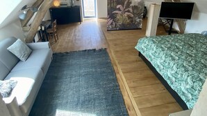 Un dormitorio cálido en el ático, con techo inclinado, vigas de madera, un sofá, una cama, un ventanal grande y un cuadro.