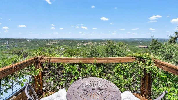 A wooden deck with a metal table and chairs, overlooking a scenic landscape with greenery and distant buildings.