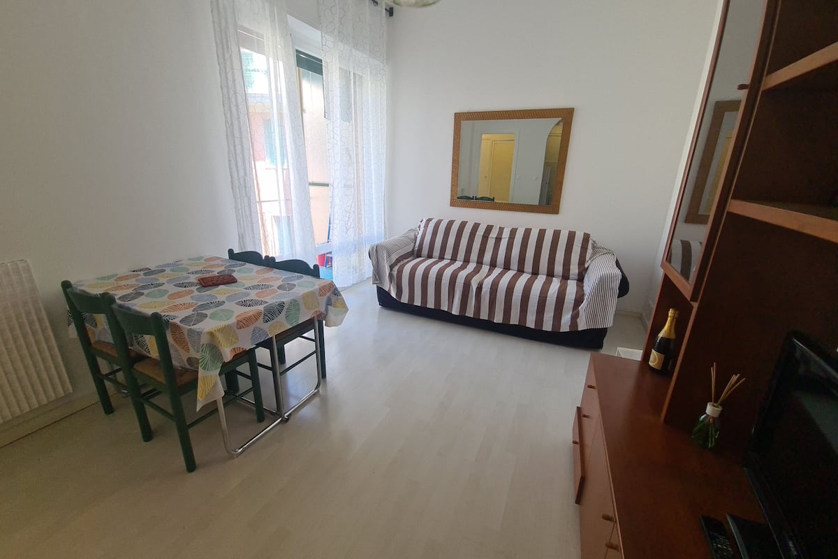 A dining area with a table and chairs, a sofa, a mirror, and a bottle on a wooden cabinet.