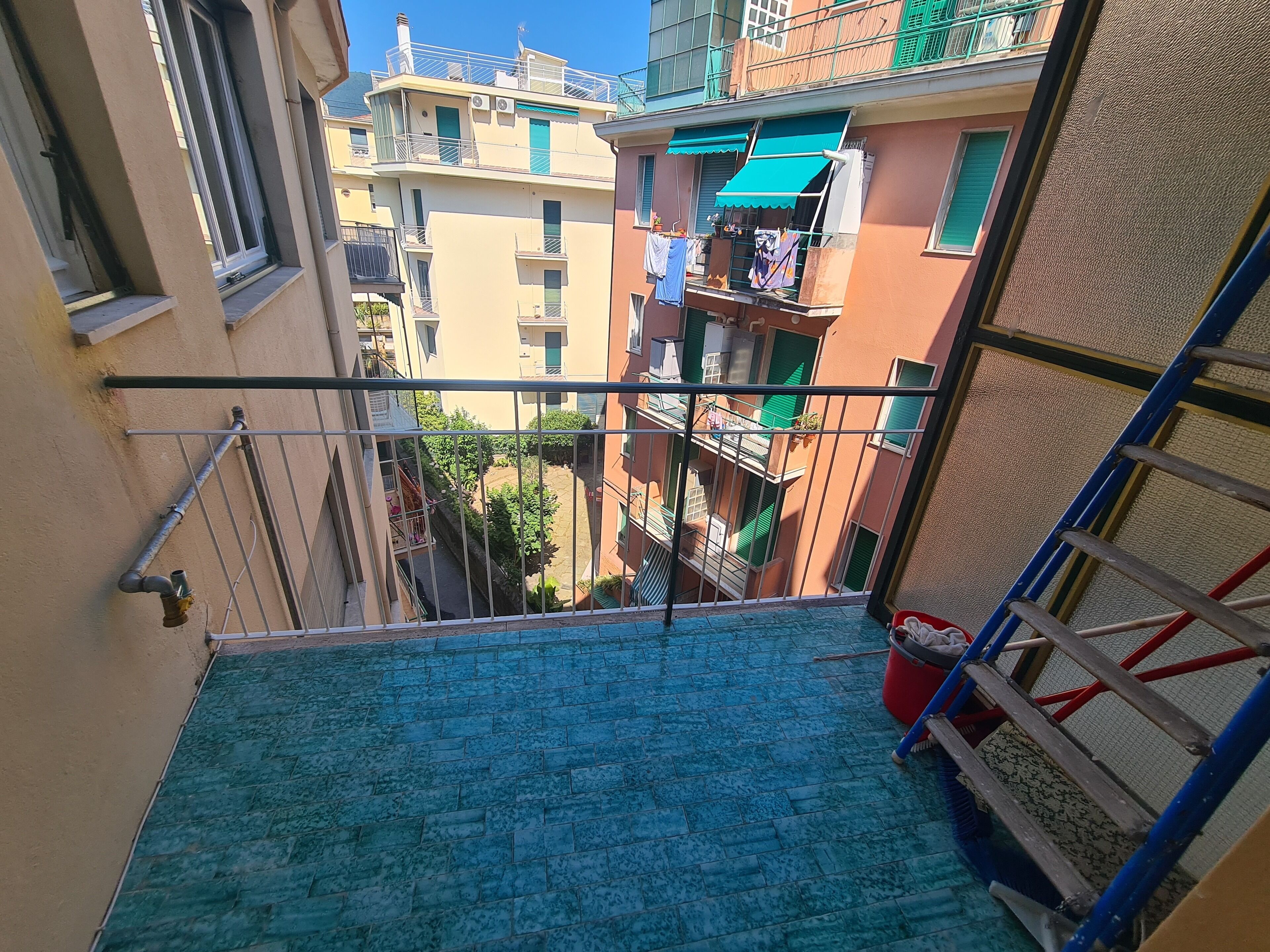 Uma escada estreita com um patamar azulejado em azul, grades de metal e um balde vermelho.