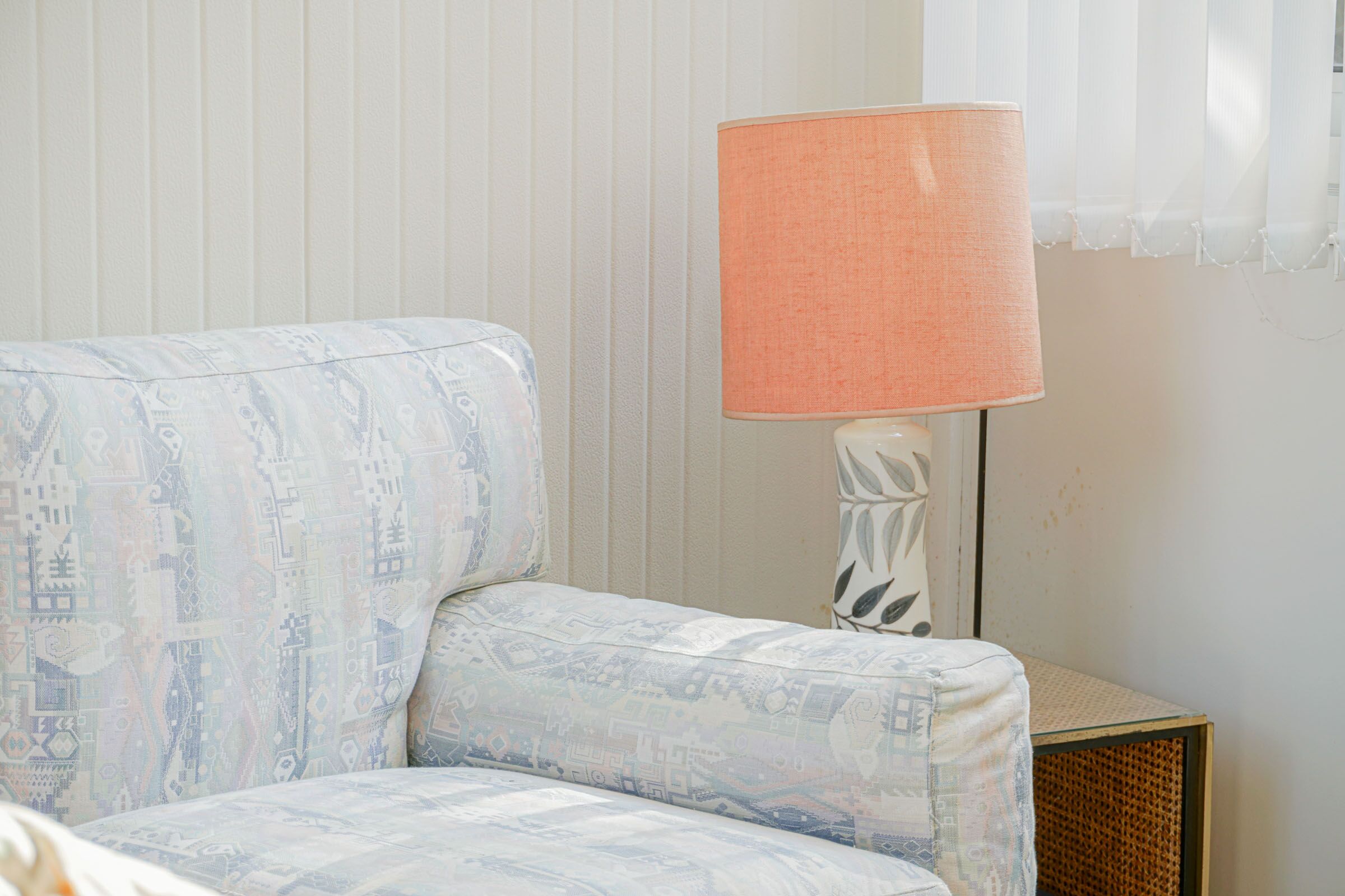 Una habitación con un sofá estampado, una lámpara con pantalla color melocotón y una mesita auxiliar de mimbre.