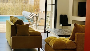 Een moderne woonkamer met een bank, een tafel en een televisie. Door de glazen deuren is een zwembad zichtbaar.