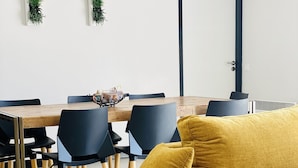 Een eetruimte met een houten tafel en zwarte stoelen, een mosterdgele bank en wandplanters.