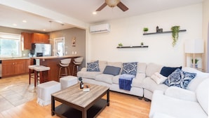 A modern living room with a sofa, coffee table, and bar stools. There is a ceiling fan, a kitchen area with cabinets and a refrigerator, and a dining table with chairs. - Cozy Boston Homestead : Sleeps 8, Pet Friendly (Framingham)