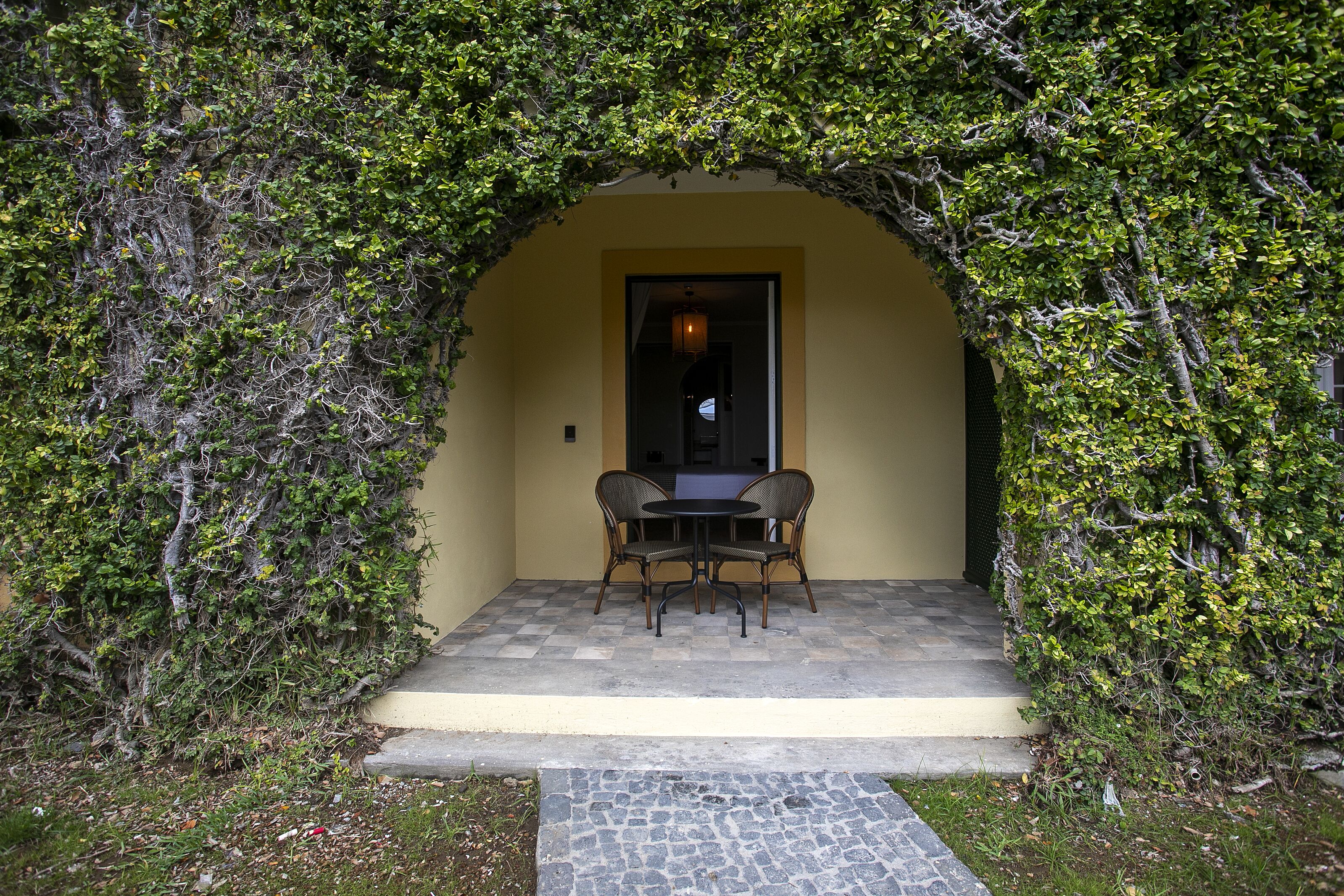 An outdoor seating area with a table and chairs under an archway of dense foliage.