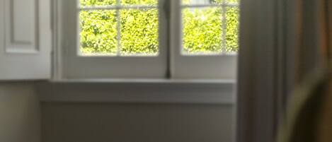 Una cama con ropa blanca, una ventana con vistas a follaje, y una cortina que cubre parcialmente la ventana.