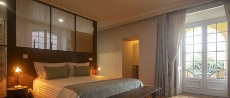 A bedroom with a bed, bedside lamps, a bench, a large window, and a view of greenery outside.