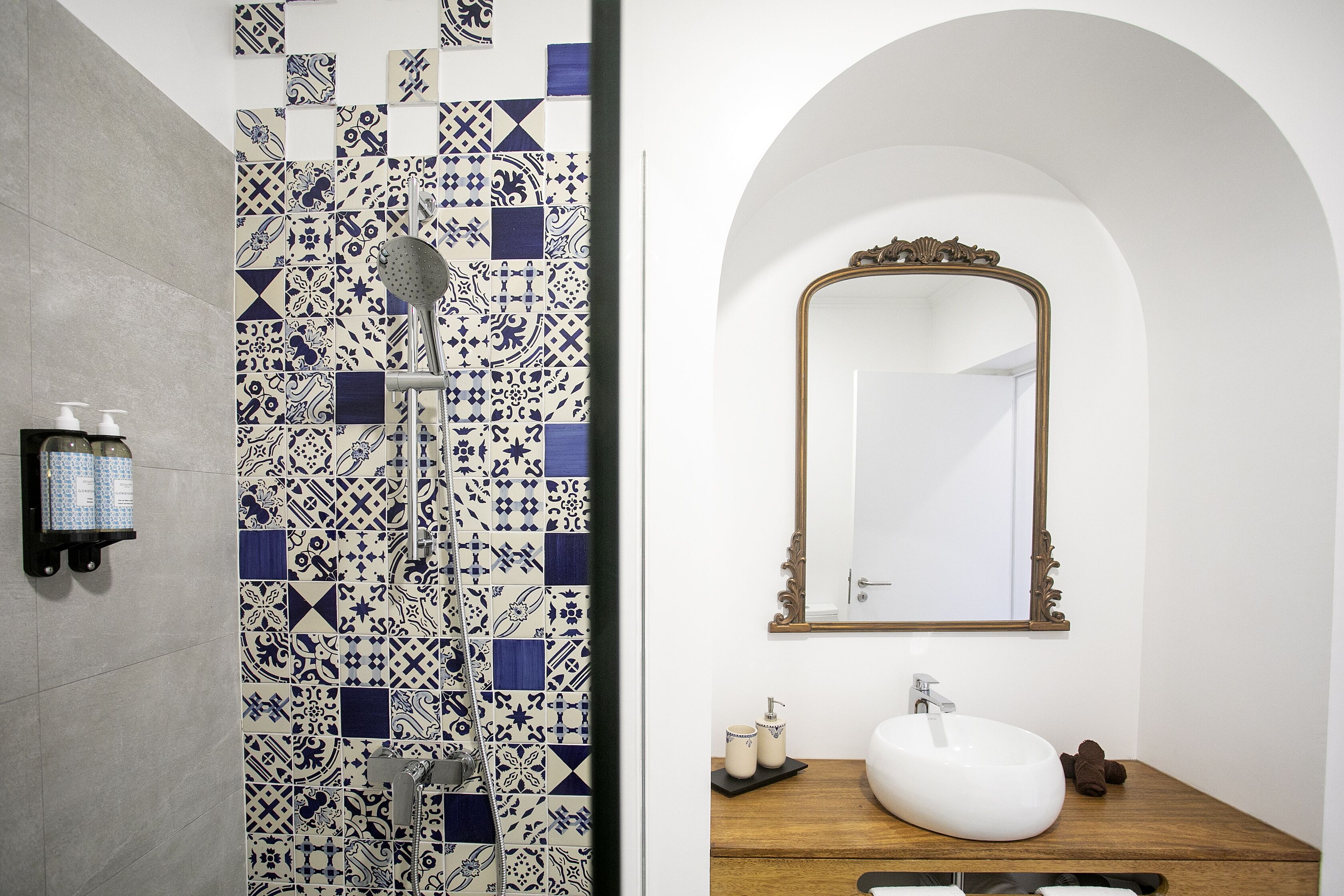 A bathroom with a tiled shower wall, a wooden vanity with a white sink, and a mirror with an ornate frame.