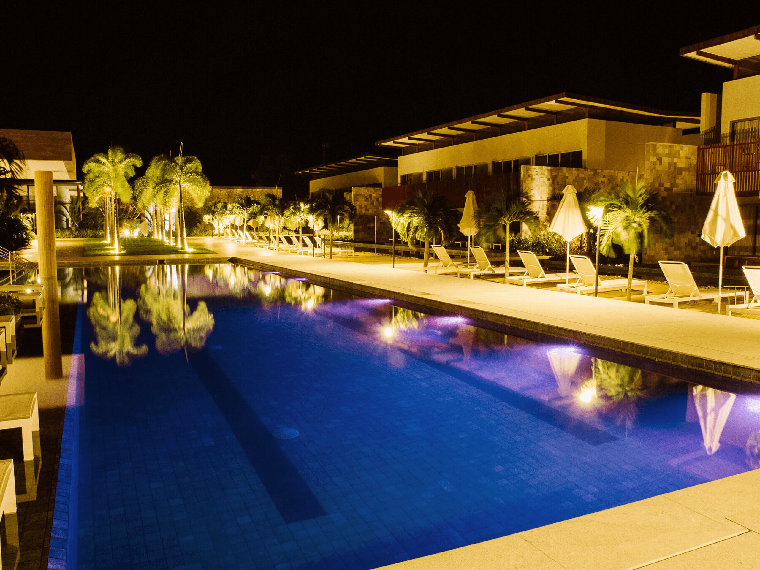 A nighttime swimming pool with lounge chairs and umbrellas, surrounded by palm trees and modern architecture.