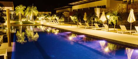 A nighttime swimming pool with lounge chairs and umbrellas, surrounded by palm trees and modern architecture.