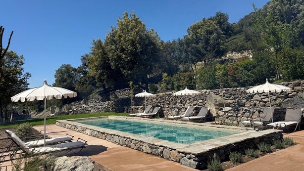 Una piscina con sillas de descanso y sombrillas, rodeada de muros de piedra y árboles.