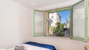 A bedroom with a bed, a window with green frames, a view of a street with buildings, and a small plant on the windowsill.