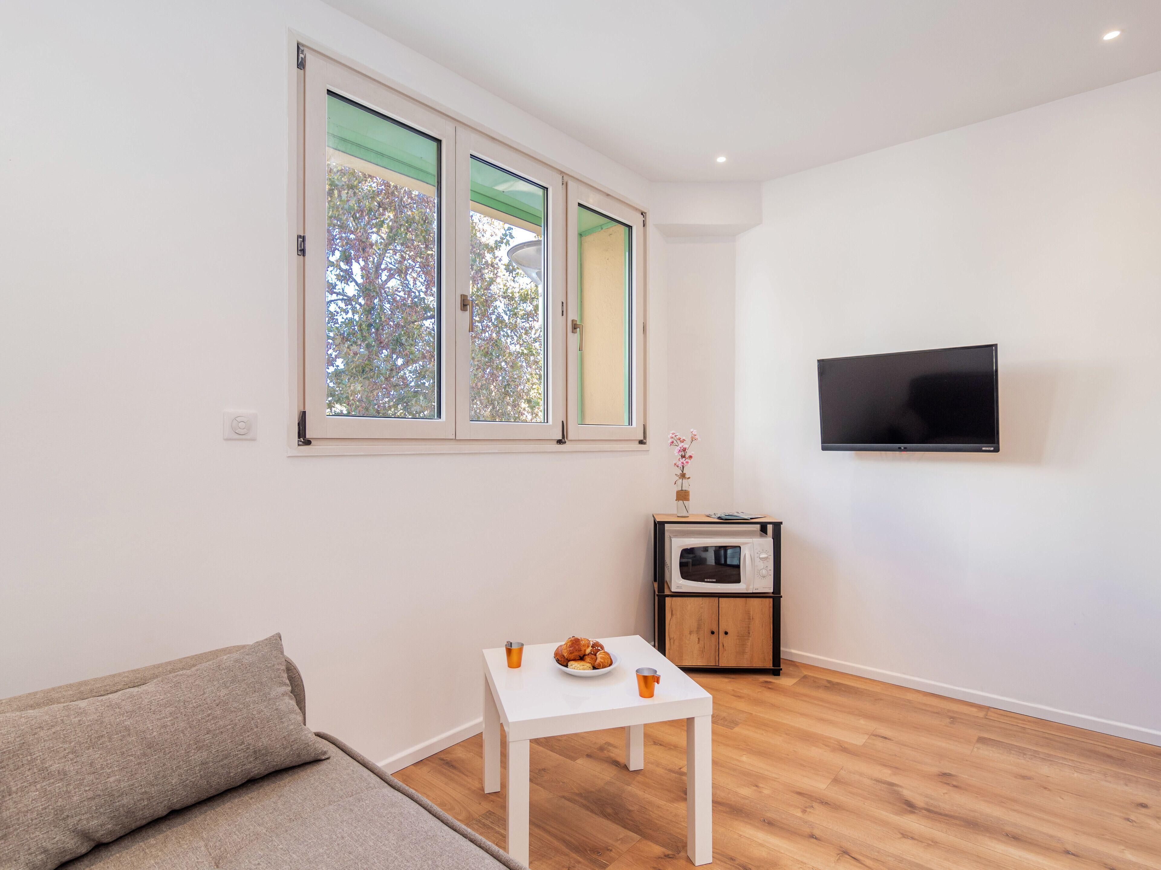 Een moderne woonkamer met een witte bank, een klein wit tafeltje met een kopje en een bord, een aan de muur bevestigde televisie, en een raam met uitzicht op bomen.