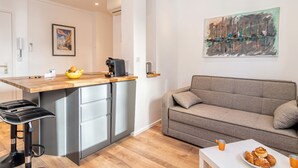 A modern kitchen with a bar stool, a dining table with pastries, and a grey sofa.