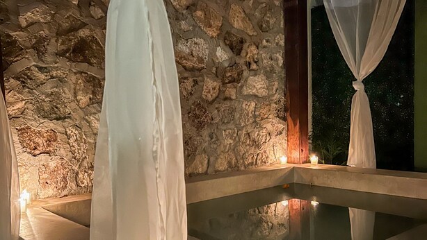 A stone-walled room with a stone bathtub, white curtains, and candles. - Hotel Maculan (Valladolid)
