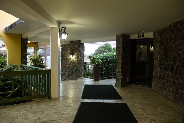 A hotel lobby with a tiled floor, a green railing, a large potted plant, and a sign indicating a room number. - Crown Point Beach Hotel (Crown Point)