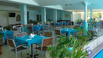 A restaurant with multiple tables and chairs, some with blue tablecloths, and potted plants around the area.