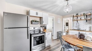 A modern kitchen with a stainless steel refrigerator, built-in oven, and white cabinetry. There is a wooden dining table with chairs and a chandelier above. - Across the Street From Von Stiehl-newly Renovated (Algoma)
