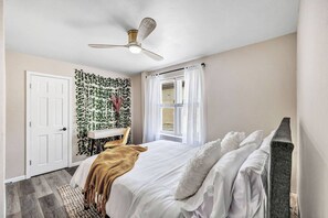 A bedroom with a bed, a ceiling fan, a window with curtains, a door, and a wall with green foliage. - Walking Distance to Everything- Newly Renovated (Algoma)