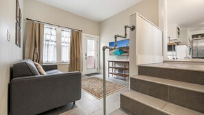 Een moderne woonkamer met een grijze bank, een trap naar een keuken, en uitzicht buiten door grote ramen.