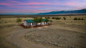 A small, single-story house with a green roof and a fenced porch, surrounded by a dry, open landscape with distant mountains.