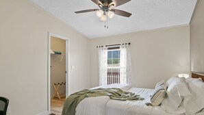A bedroom with a bed, a ceiling fan, a window with curtains, a door leading to a bathroom, and a small table.