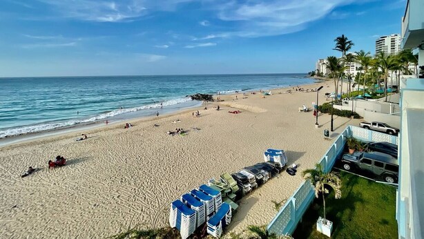 A beach with a clear sky, a sandy shore, and a few people. There are buildings in the background and a few parked cars.