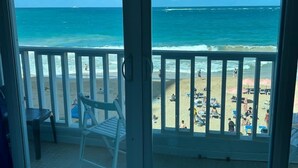 A room with a sliding glass door leading to a balcony overlooking a beach with people and umbrellas.