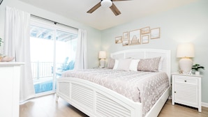Une chambre à coucher comprenant un lit, un ventilateur de plafond, une table de chevet, une lampe et une fenêtre avec des rideaux.