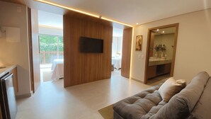 A modern living room with a sofa, a flat-screen TV mounted on a wooden wall, and a dining area visible through an open doorway. - Flat Ilê de Pipa Luxxor - By Almare (Tibau do Sul)