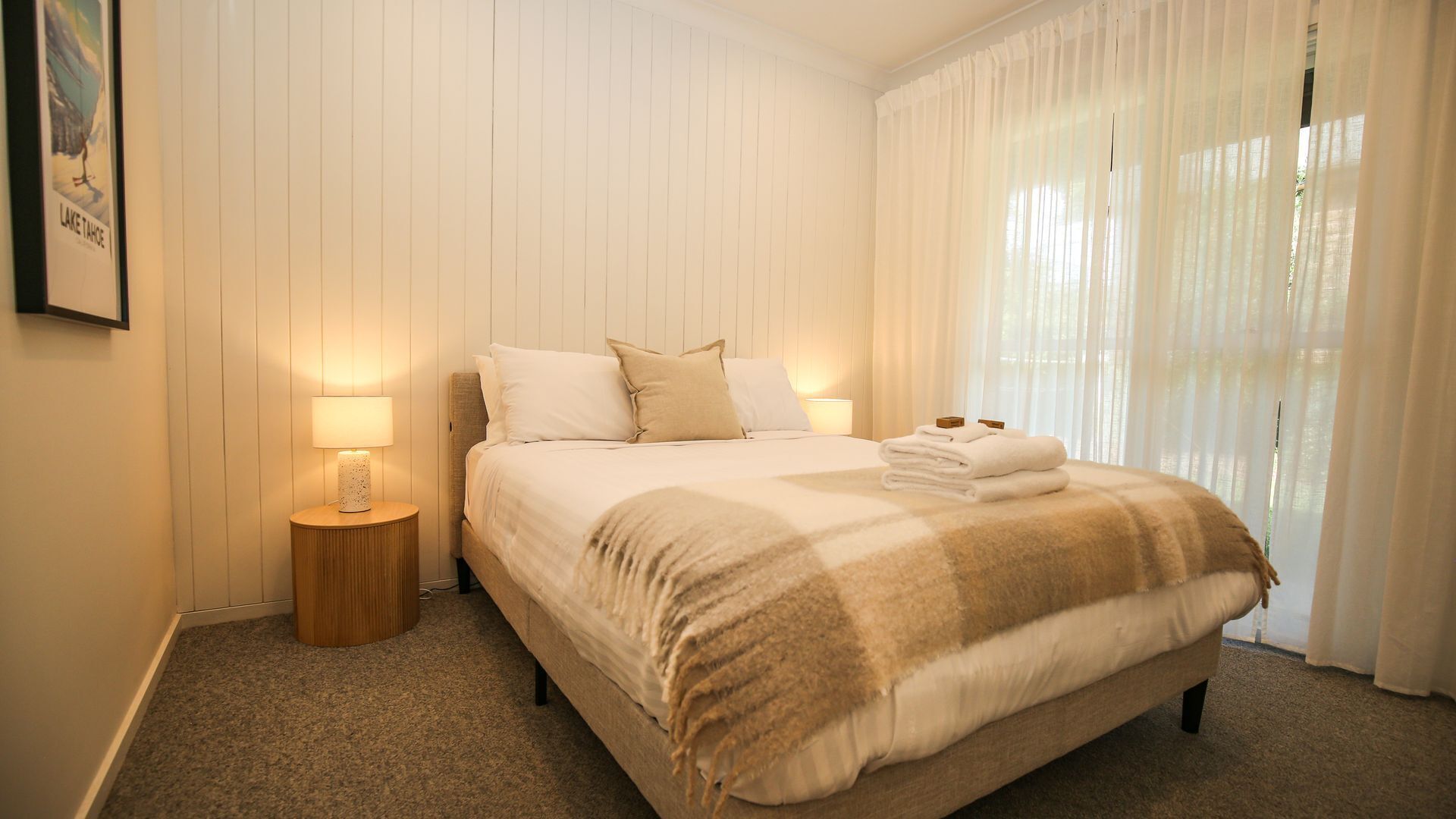 A neatly made bed with a blanket, two bedside lamps, and a window with sheer curtains.