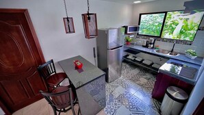 Cuina compacta amb taula de dinar, cadires, nevera, microones i un finestra gran que mostra vegetació.