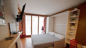 Quarto com cama, escrivaninha, cadeira, televisão na parede e janela com cortinas.