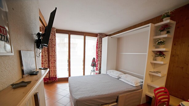 Een slaapkamer met een bed, een bureau, een stoel, een aan de muur bevestigde televisie en een raam met gordijnen.
