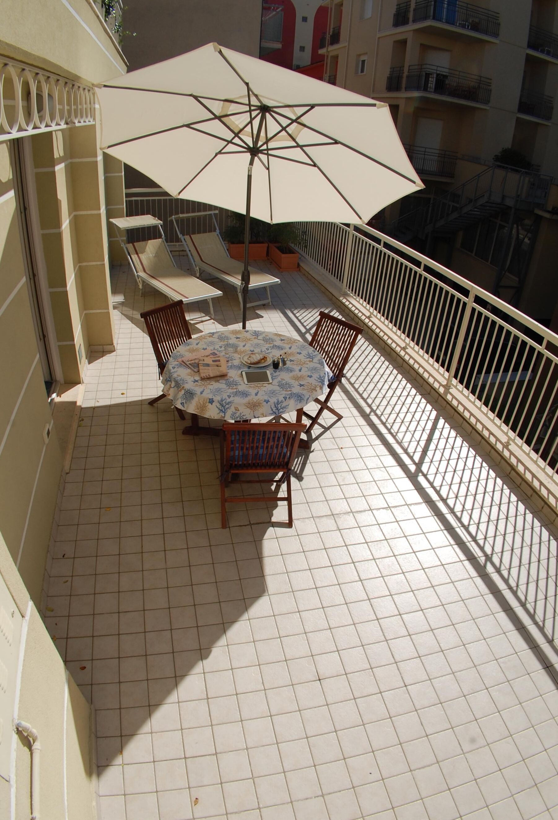 Un balcon avec un sol carrelé, une table ronde entourée de chaises et un grand parasol.