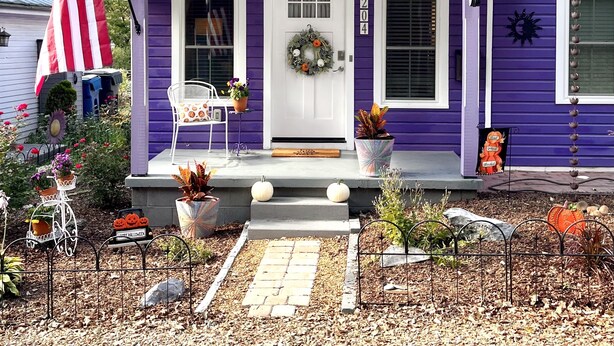 Een paars huis met een witte deur, een veranda en een voortuin met planten en pompoenen.