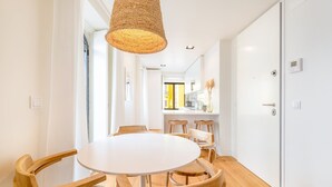 Un comedor moderno con una mesa redonda y sillas de madera, una lámpara colgante y vistas a una cocina con armarios blancos y un ventanal.
