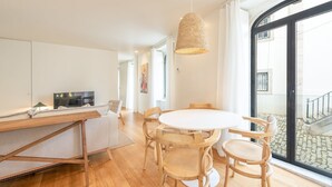 Una sala de estar moderna con una mesa redonda de madera y sillas, un televisor sobre un mueble blanco, y un ventanal con vistas a una calle adoquinada.