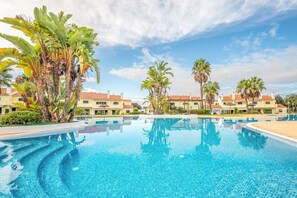 Una piscina con bordi piastrellati, circondata da palme e da una fila di edifici con tetti rossi.