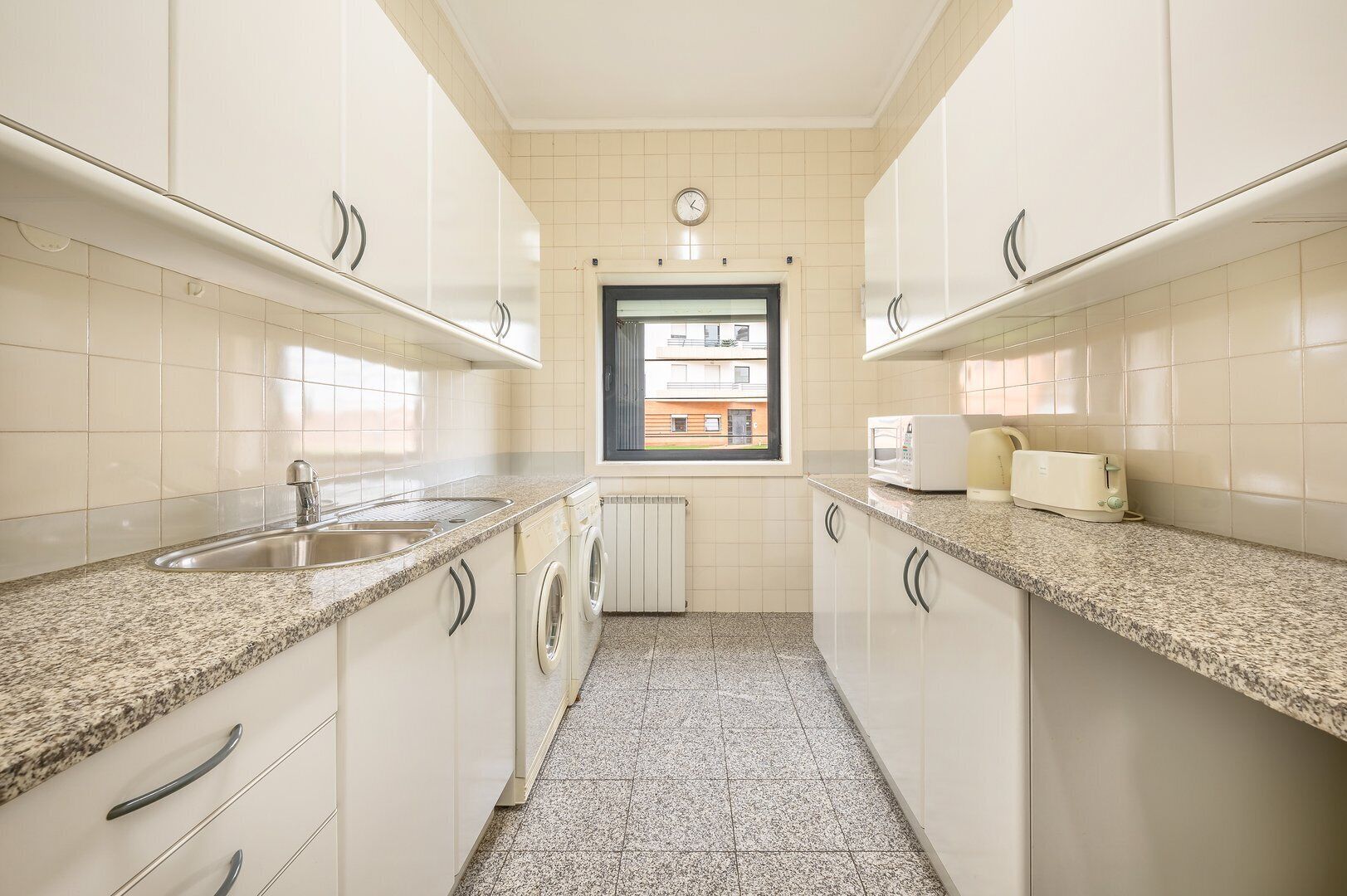 A narrow kitchen with white cabinets, a granite countertop, a built-in washing machine, and a window with a view of buildings.