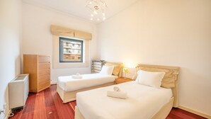A hotel room with two beds, a wooden dresser, a radiator, and a window with a view of buildings.