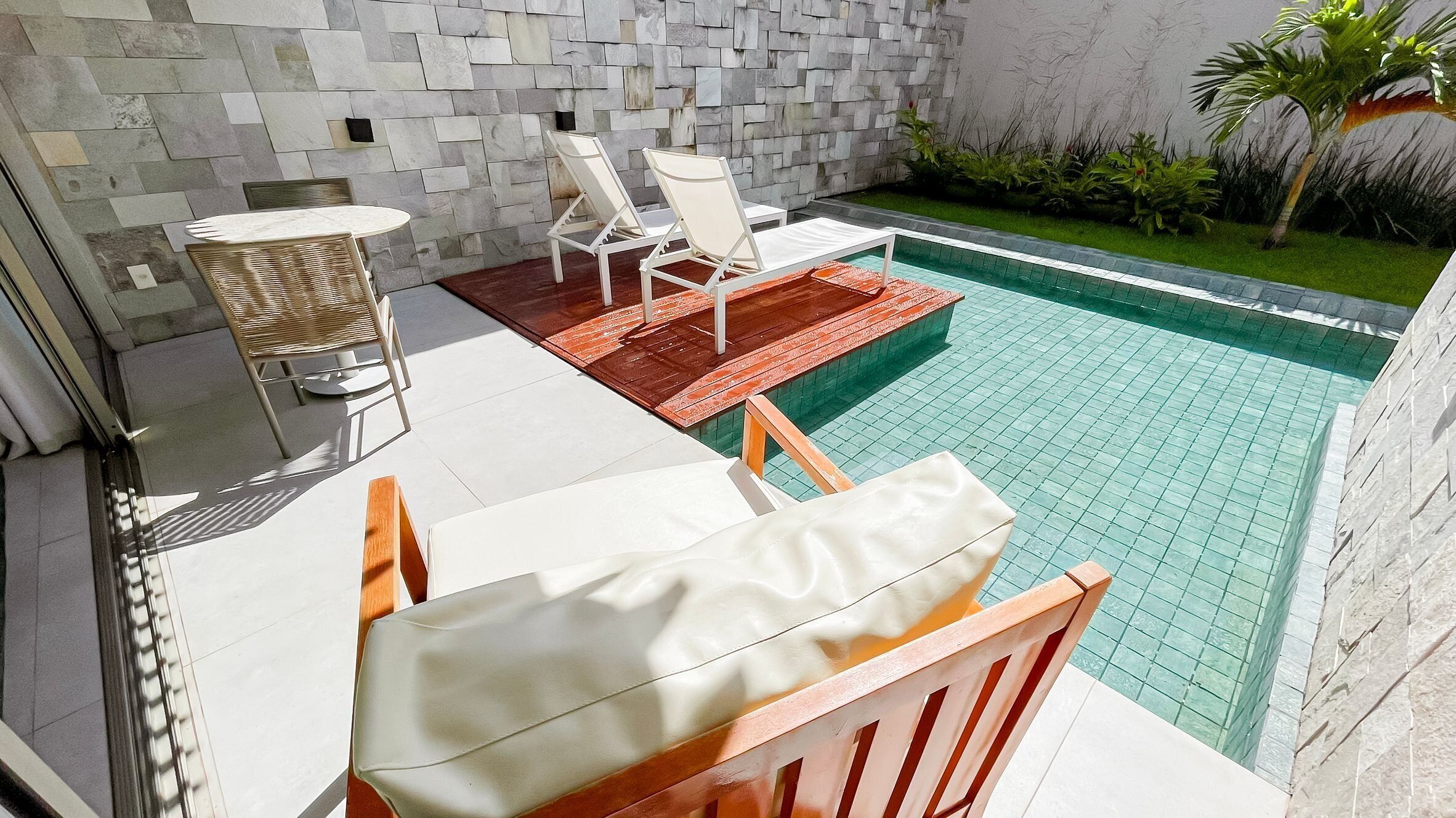 Zona junto a la piscina con pared de piedra, cubierta de madera y sillones blancos.