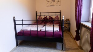 A single bed with a metal frame, a purple bedspread, and several pillows. There is a radiator on the right and a window with purple curtains.
