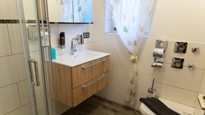 A bathroom with a wooden vanity, a glass shower enclosure, a toilet, and a window with a floral curtain.